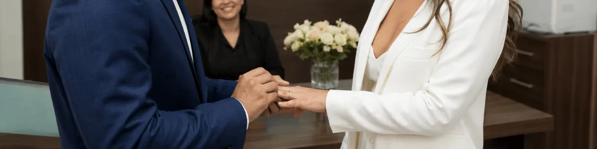 Casal sorridente troca alianças em um cartório moderno, com uma funcionária ao fundo acompanhando a cerimônia em um ambiente discreto, institucional e acolhedor