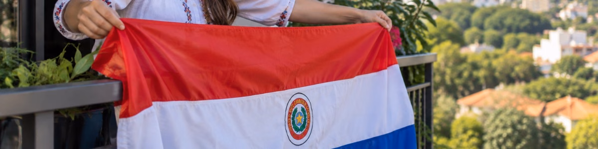 Mulher sorridente estendendo a bandeira do Paraguai na varanda de sua residência, em um ambiente iluminado pela luz natural, com prédios e vegetação ao fundo.