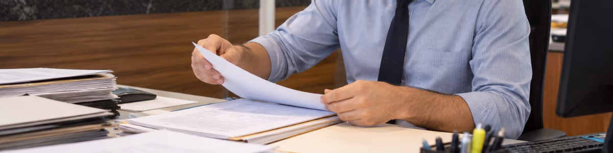 Funcionário de cartório organizando e conferindo documentos em sua mesa, em um ambiente profissional, com arquivos e outros atendentes ao fundo